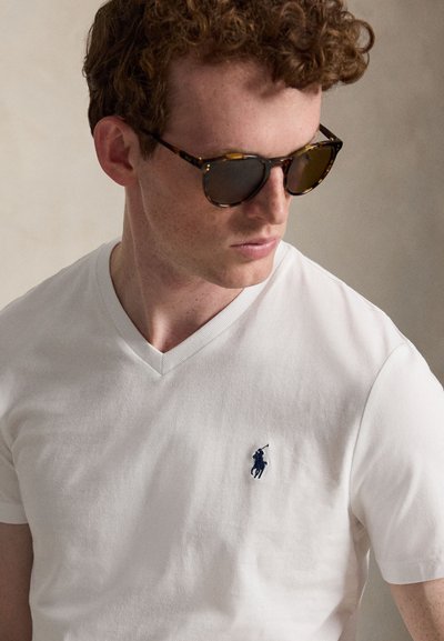 Muž s kučeravými červenými vlasmi, ktorý nosí slnečné okuliare so želvovinovým rámom a biele tričko s výstrihom do V s malým tmavomodrým logom hráča póla.