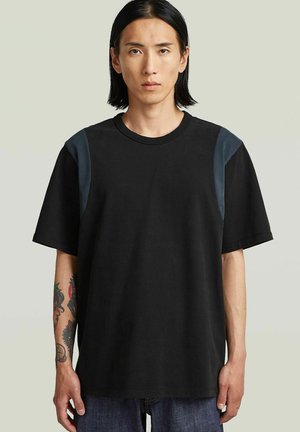 Jeune personne aux longs cheveux noirs et bras droit tatoué, portant un t-shirt noir ample avec des taches foncées sur les épaules, sur fond uni.