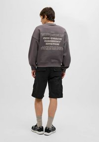 Sweatshirt gris surdimensionné avec du texte dans le dos, associé à un short cargo noir, des chaussettes beiges et des baskets noires. Tenue simple et décontractée.