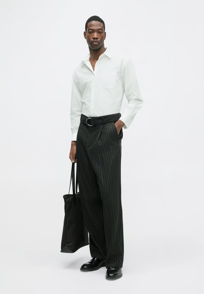Witte overhemd met lange mouwen en een zak, zwarte gestreepte broek, zwarte riem en zwarte tote bag. Model draagt gepolijste zwarte schoenen.
