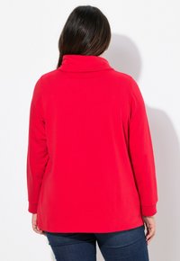 Maglietta rossa a maniche lunghe con colletto alto e texture morbida. Presenta una vestibilità rilassata e un orlo che cade sotto i fianchi. Nessuna chiusura visibile.