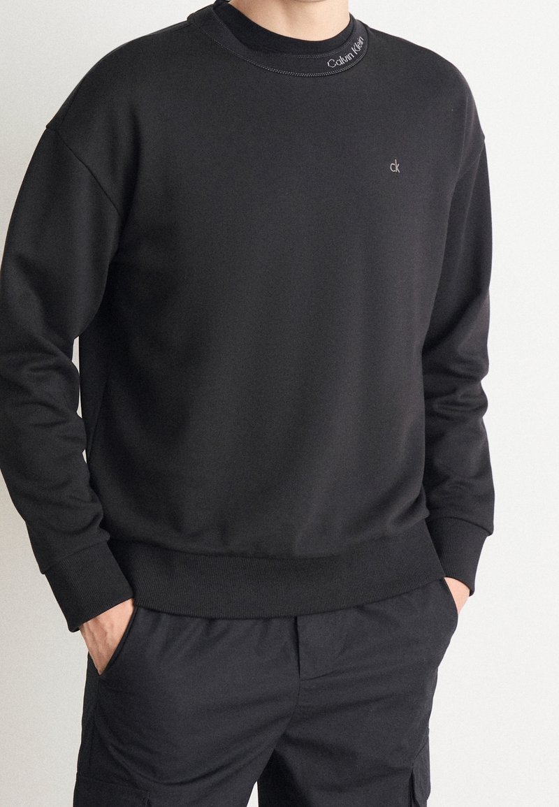 Personne portant un sweat-shirt noir Calvin Klein avec un petit logo sur la poitrine et le col, les mains dans les poches, sur un fond clair uni.