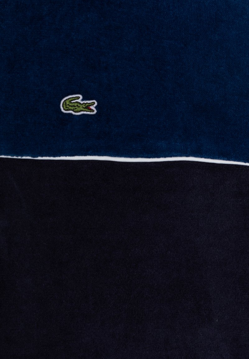 Tissu en coton marine avec une partie inférieure bleu foncé. Présente un petit logo crocodile vert brodé. Texture lisse avec une bordure blanche.