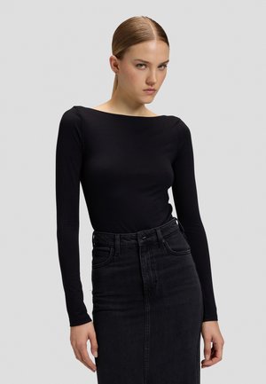 Jeune femme aux cheveux châtain clair lisses attachés, portant un haut noir moulant à manches longues et une jupe en jean noire taille haute sur fond gris.