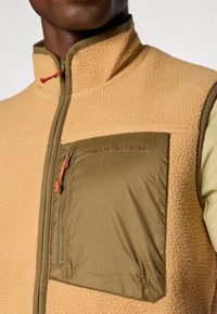 Fleece vest in licht bruin met een bruine accent. Kenmerkt zich door een hoge kraag, een met rits afsluitbare borstzak en een gestructureerde stof. Merklogo geborduurd op de borst.