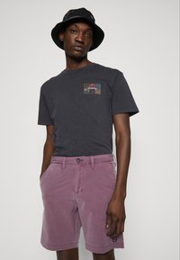 Mies, który nosi czarny kapelusz z rondem, ciemnoszary T-shirt z małym wielokolorowym wzorem oraz szorty w kolorze mauve z kieszeniami, stoi na tle białego tła.