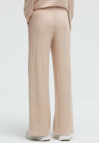 Beige gerippte, weit geschnittene Hosen aus weichem Stoff mit strukturierter Oberfläche und entspanntem Sitz, kombiniert mit weißen Sneakers.