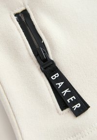 Cremefarbener Stoff mit einem schwarzen Reißverschluss. Ein schwarzes gewebtes Etikett mit "BAKER" ist in der Nähe des Reißverschlusses angebracht. Die Textur ist glatt, mit sichtbaren Nähten.