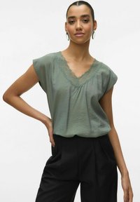 Blusa verde a maniche corte con scollo a V e rifiniture in pizzo. Tessuto morbido, vestibilità rilassata, abbinata a pantaloni neri a vita alta.