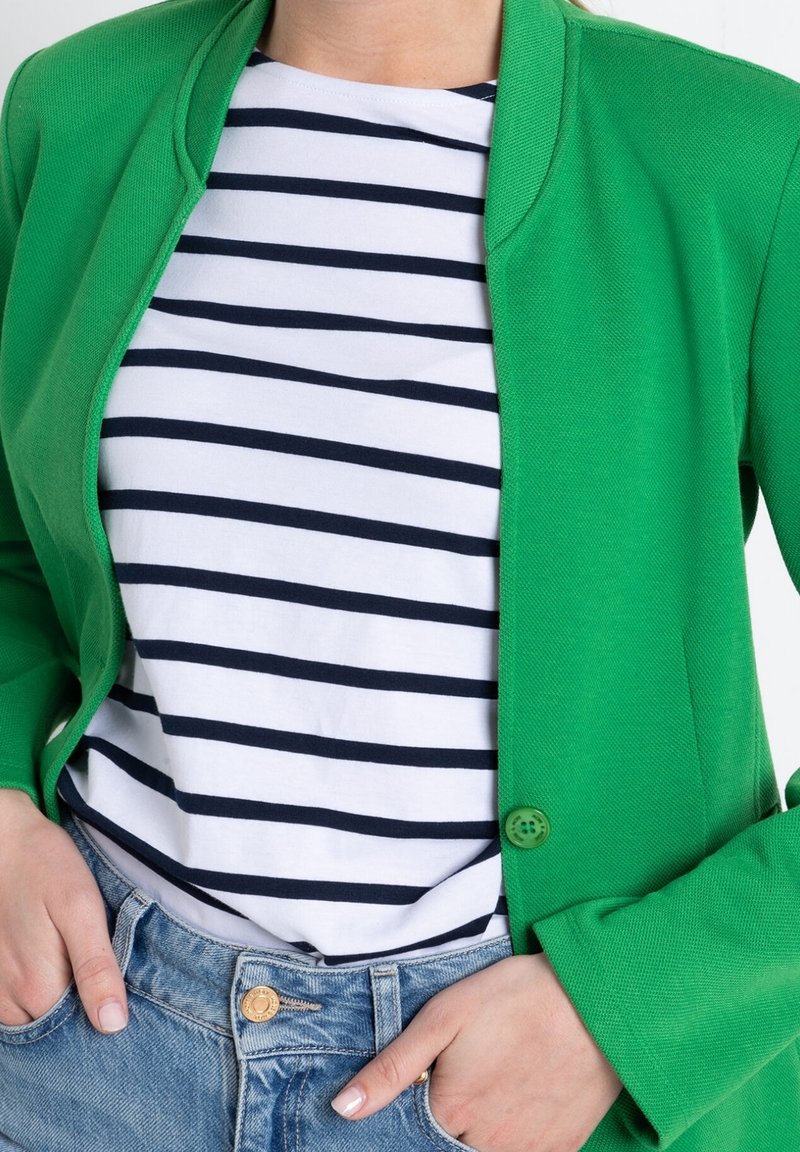 Grüner strukturierter Blazer mit rundem Kragen und einem Knopfverschluss, getragen über einem weiß-blau gestreiften Hemd. Hellblaue Jeans sind sichtbar.