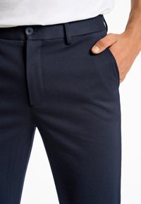 Pantalons ajustés bleu marine en tissu lisse, avec fermeture par bouton et poches latérales ; conçus avec une coupe slim et des lignes épurées.