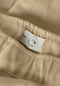 Beige fabric with a textured weave. Features a white elastic band with a button closure and a tag stitched into a seam.