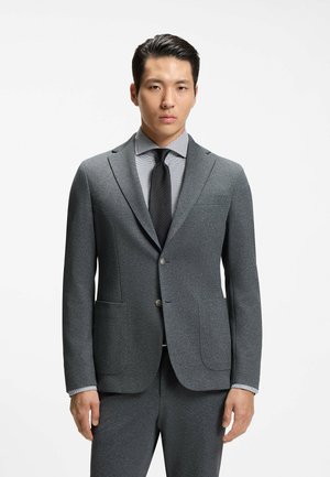 Hombre vistiendo un traje gris con camisa de cuadros y corbata negra, de pie frente a un fondo blanco liso.