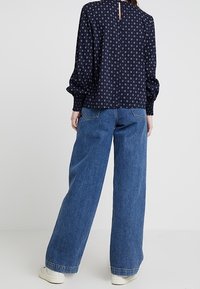 Blusa blu con un design a fantasia e maniche a sbuffo, abbinata a jeans in denim blu larghi e sneakers bianche. Vista posteriore visibile.