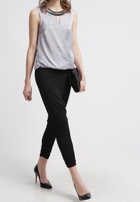 Szary bezrękawnik z dekoltem w kształcie łezki i koralikowym wykończeniem, w połączeniu z dopasowanymi czarnymi spodniami i czarnymi szpilkami, utrzymywany w ręku przez kopertówkę.