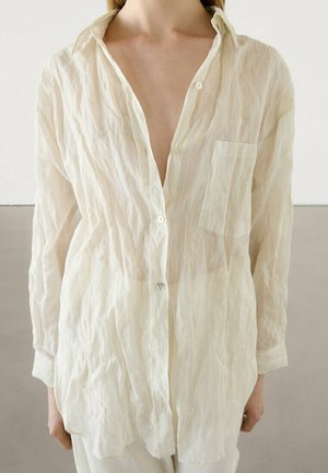 Person wearing a loose, long-sleeve, off-white wrinkled button-up shirt with an open collar and chest pocket, standing against a neutral background.
