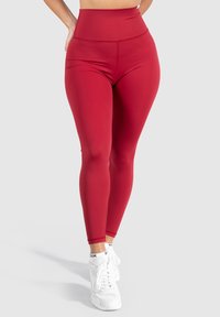 Leggings rojos de talle alto fabricados en una tela elástica, con una textura suave y un ajuste entallado, combinados con zapatillas deportivas blancas.