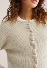 Cardigan bej cu dungi, realizat din lână texturată, având o fâșie cu nasturi volantați colorați și un guler rotund.