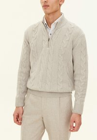 Light gray cable-knit sweater with a high collar, zip closure, and ribbed cuffs. Worn over a collared shirt and paired with matching trousers.