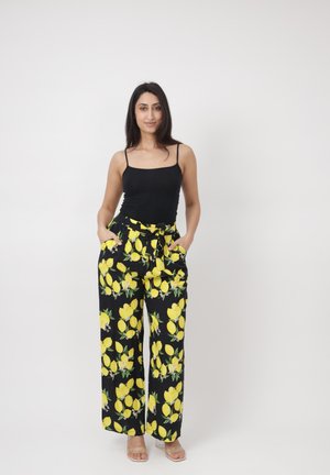 Femme debout les mains dans les poches, portant un débardeur noir et un pantalon large imprimé de citrons jaunes sur fond noir.