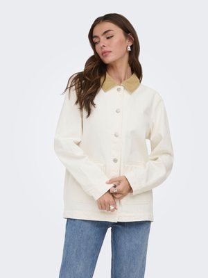 Kvinde iført en cremefarvet jakke med knapper og en beige krave, iført blå jeans, kigger ned med hænderne foldet foran sig.