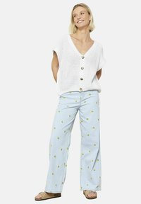 Cardigan bianco lavorato a maglia con maniche corte e bottoni, abbinato a pantaloni ampi blu chiaro con motivi di banane gialle. Inclusi calzature casual.