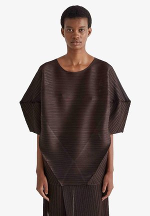 Persona snella indossa un top marrone scuro plissettato con maniche larghe e design angolare e geometrico, in piedi su sfondo bianco uniforme.