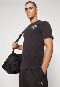 Black T-shirt with faded texture and white Puma logo. Worn with black pants and holding a black sports bag with mesh accents.