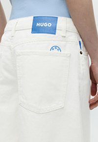 Κοντινή λήψη πίσω τσέπης από λευκό τζιν με μπλε ετικέτα «HUGO» και κυκλικό λογότυπο, φορεμένη με ανοιχτό μπλε τοπ.