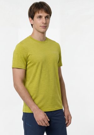 Giovane uomo in piedi, che indossa una maglietta gialla a maniche corte e pantaloni blu scuro, rivolto leggermente verso sinistra su uno sfondo bianco.