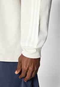 A camisa branca de mangas longas apresenta punhos canelados, três riscas verticais ao longo do braço e um tecido texturizado, combinada com calças azul-escuro.