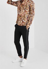 Langärmliges Hemd aus einem mehrfarbigen, gemusterten Stoff mit einem kragenförmigen Ausschnitt, kombiniert mit schwarzen Jogginghosen mit weißen Seitenstreifen und weißen Turnschuhen.