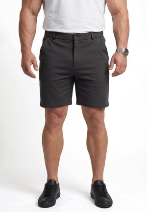 Mand iført sorte skræddersyede shorts, hvid t-shirt, sorte sneakers og et sølvfarvet armbåndsur, stående med arme afslappet ned langs siderne.