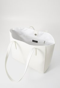Hvid tote bag lavet af tekstureret materiale, med lange håndtag og tryklås. Indeholder en lynlåslomme indeni.