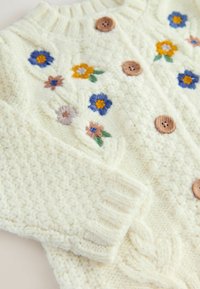 Cardigan tricotat alb, decorat cu flori brodate în galben, albastru și roz. Nasturi de lemn și manșete tricotate oferă textură și detaliu.