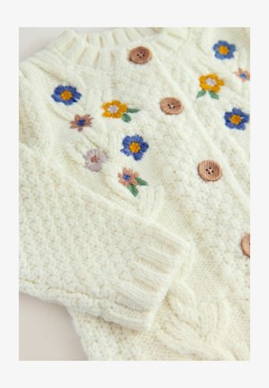 Biely pletený sveter s vyšívanými kvetmi v žltej, modrej a ružovej farbe. Drevené gombíky a rebrované manžety dodávajú textúru a detaily.