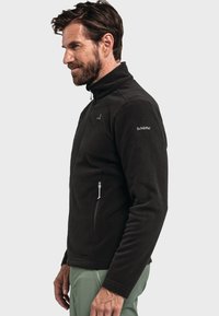 Chaqueta de forro polar negra con cuello alto, cremallera frontal y bolsillos laterales; presenta un logotipo en el hombro izquierdo y una textura suave y lisa.