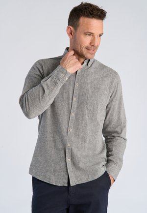 Mand iført en grå, langærmet skjorte med knapper, med den ene hånd ved halsen og den anden i lommen.