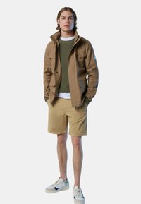 Bruine ritssluiting jack, groene gebreide trui, witte T-shirt, kaki shorts en witte sneakers met groene accenten. Eenvoudig ontwerp, minimale details.