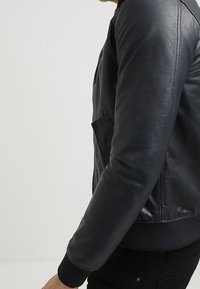 Svart läderbomberjacka med ribbade manschetter och fåll, med en dragkedja fram och sidoficka. Slät textur och figursydd design.