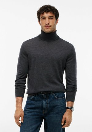Mann mit dunklem lockigem Haar, der einen dunkelgrauen Rollkragenpullover, blaue Jeans und eine schwarze Armbanduhr trägt und vor einem weißen Hintergrund steht.