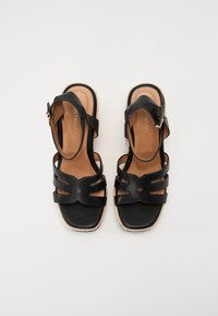 Sandales en cuir noir avec plusieurs lanières croisées, une semelle intérieure rembourrée de couleur beige et une semelle extérieure en jute. Sangle de cheville réglable avec boucle.