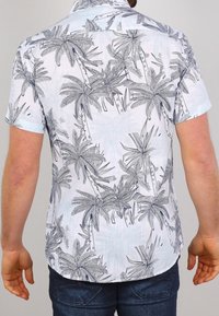 Mies joka käyttää valkoista lyhythihaista paitaa, jossa on sinisiä ja harmaita palmu kuvioita, yhdistettynä tummanssinisiin farkkuihin, kuvattuna takaa.