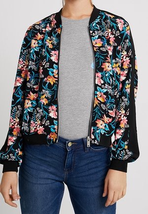 Vrouw draagt een zwarte bomberjas met kleurrijke bloemenpatronen, een grijs T-shirt en blauwe jeans, staand met ontspannen armen langs het lichaam.