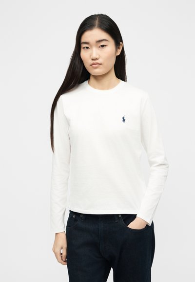 Νεαρή γυναίκα με μακριά μαύρα μαλλιά, φορώντας λευκό πουκάμισο με μακριά μανίκια και μικρό ναυτικό λογότυπο, καθώς και σκούρο μπλε παντελόνι, με το χέρι στη τσέπη.