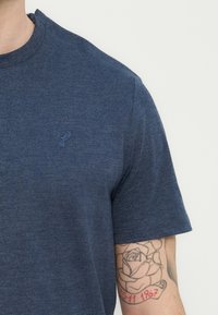 Άνδρας που φοράει μπλε μπλουζάκι με κεντητό λογότυπο πτηνού και τατουάζ τριαντάφυλλου στο πάνω μέρος του μπράτσου με ημερομηνία σε κόκκινο μελάνι.