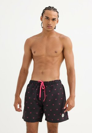 Hombre sin camiseta con el pelo trenzado, lleva pantalones cortos de baño negros con patrones de cangrejo rosa y cordón rosa, posando contra un fondo blanco.