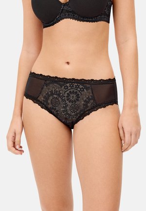 Mujer con sujetador de encaje negro y braguitas a juego de encaje floral y malla, mostrada desde la mitad del torso hasta la parte superior de los muslos.