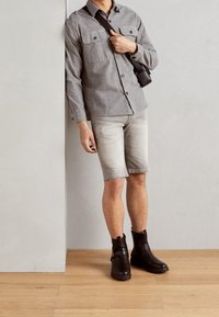 Camicia grigia a maniche lunghe con due tasche sul petto, pantaloni corti in denim chiaro e stivali neri alla caviglia con cinturini, su sfondo semplice.
