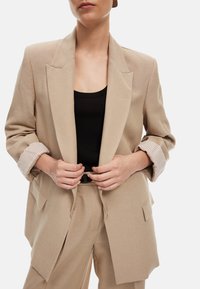 Beige räätälöity bleiseri, jossa on lovetut kaulukset, käärityt hihat, joissa on raidallinen vuori, ja kaksi etutaskua, yhdistetty mustan paidan kanssa.